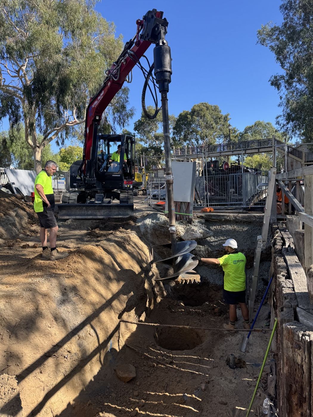 Total Piling excavator trenching along a boundary while the crew sets screw piles